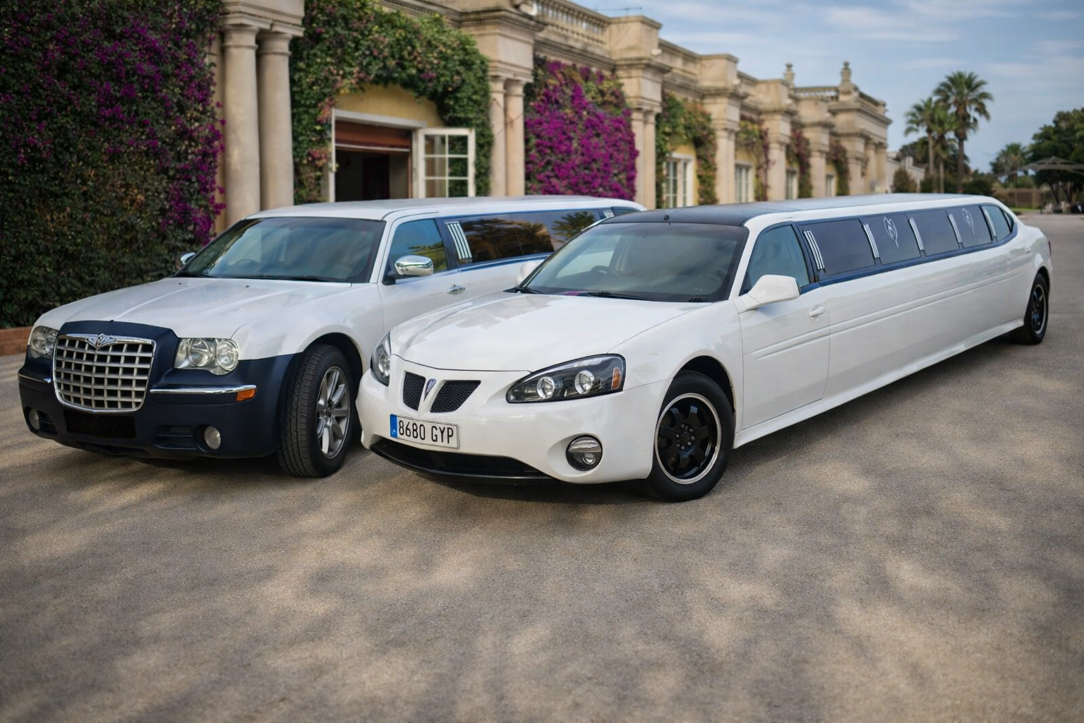 "Dos limusinas blancas estacionadas juntas frente a una elegante mansión de estilo mediterráneo con columnas y buganvillas rosas, en una fotografía realista tomada a plena luz del día.